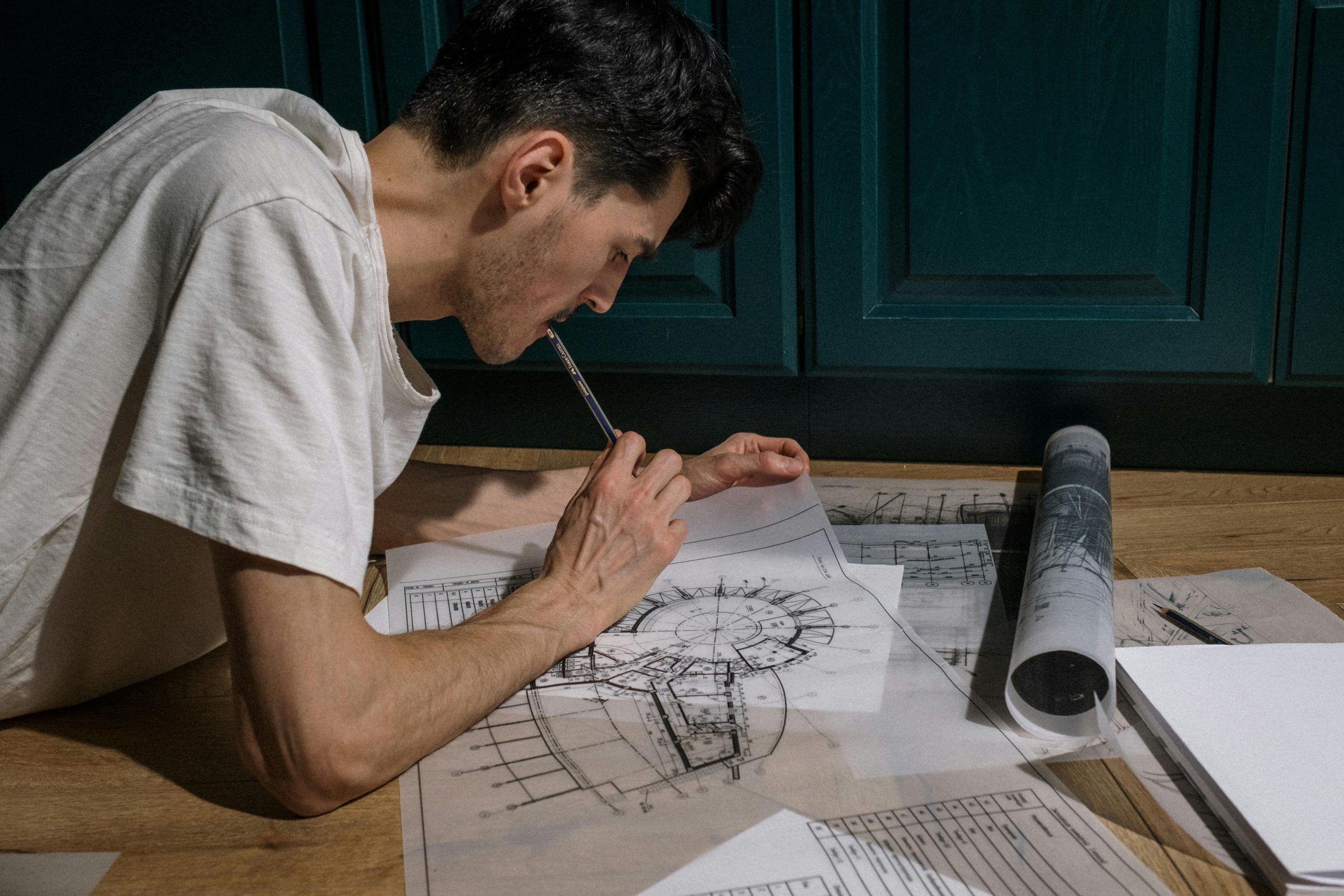 A designer laying on a wooden floor holding a pencil in his mouth while reviewing detailed architectural floor plans and structural blueprints, demonstrating meticulous joinery drafting preparation.