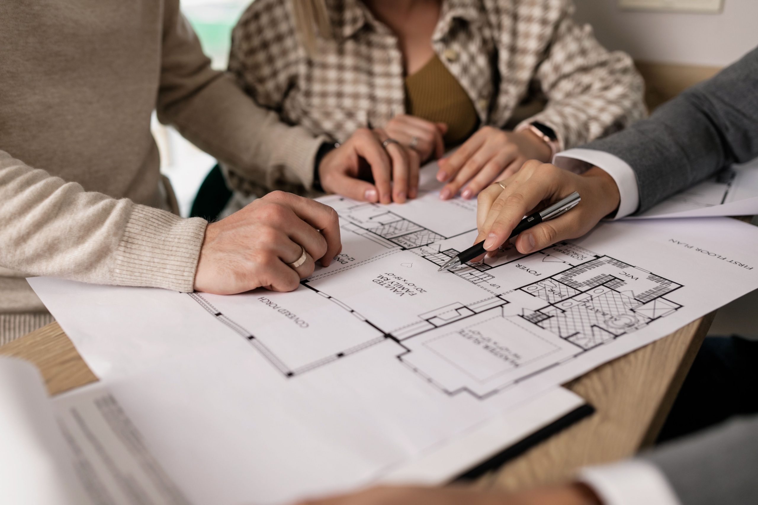 A professional consultant pointing with a pen to a first floor plan layout during a review session with clients, showcasing the initial first draft design stage.