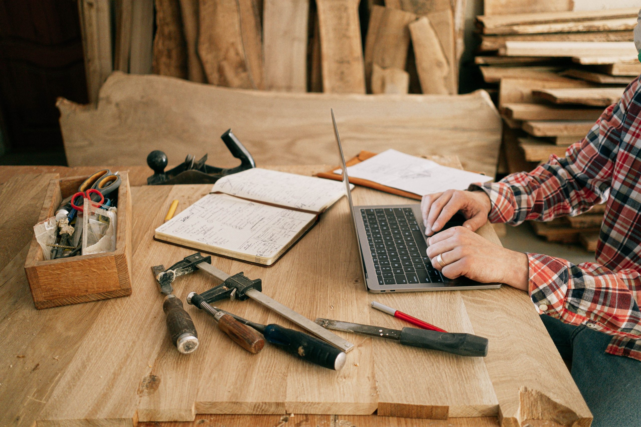 A designer using a laptop at a workbench with blueprints and woodworking tools, highlighting design consulting services.