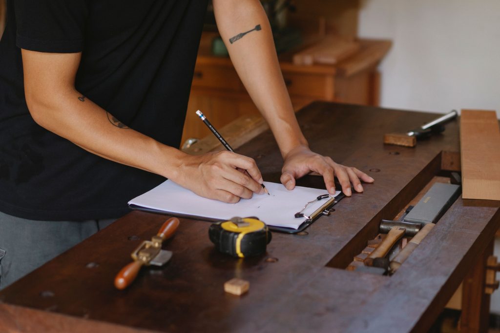 Woodworker drafting specifications on a clipboard at a workbench during the initial phase of the joinery design process.