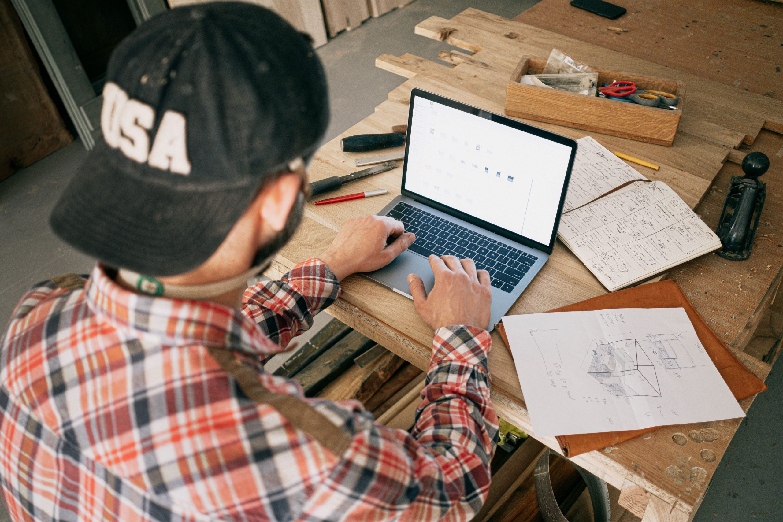 Woodworker working on a laptop surrounded by hand-drawn sketches and tools to model 3D CAD software for joinery.